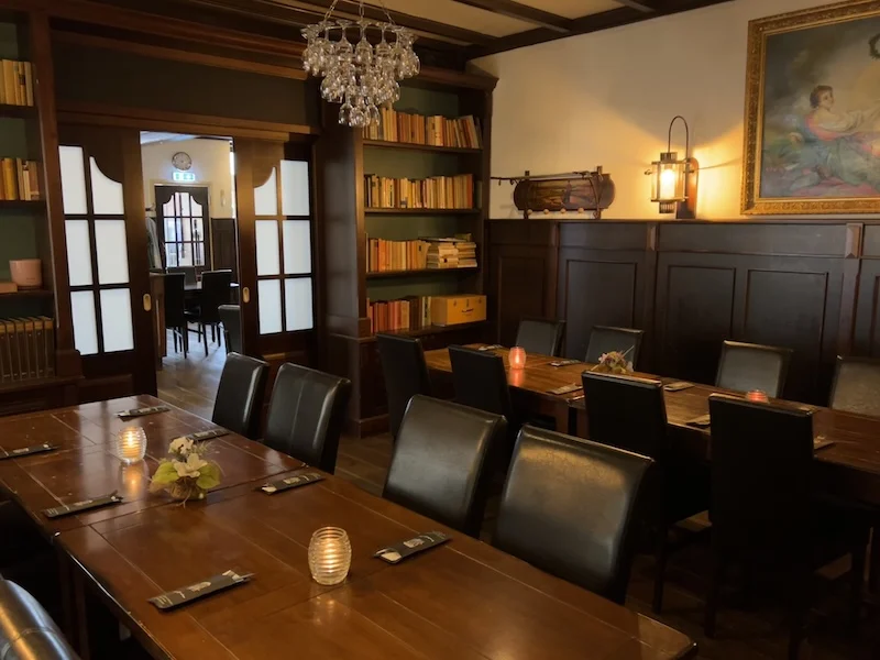 De Luifel Ruurlo – interieur met lange tafel Lange houten tafel, donkere stoelen en boekenkast in De Luifel in Ruurlo